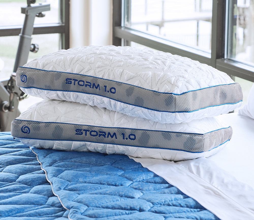 Two pillows labeled 'STORM 1.0' stacked on a blue quilted surface with a window in the background.