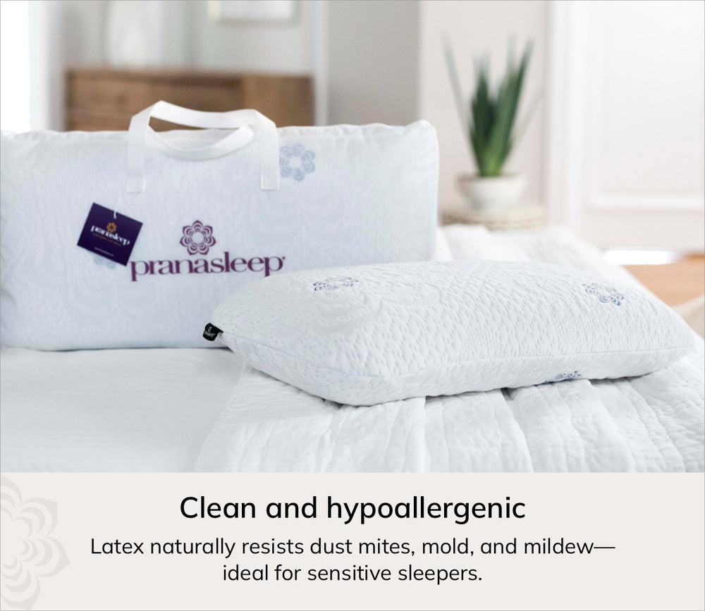 A white Pranasleep latex pillow resting on a white quilted bedspread, with the matching white pillowcase and carry bag displayed behind it. The scene is set in a softly lit bedroom with a plant in the background, and a text banner at the bottom reads 'Clean and hypoallergenic' with additional latex benefits.