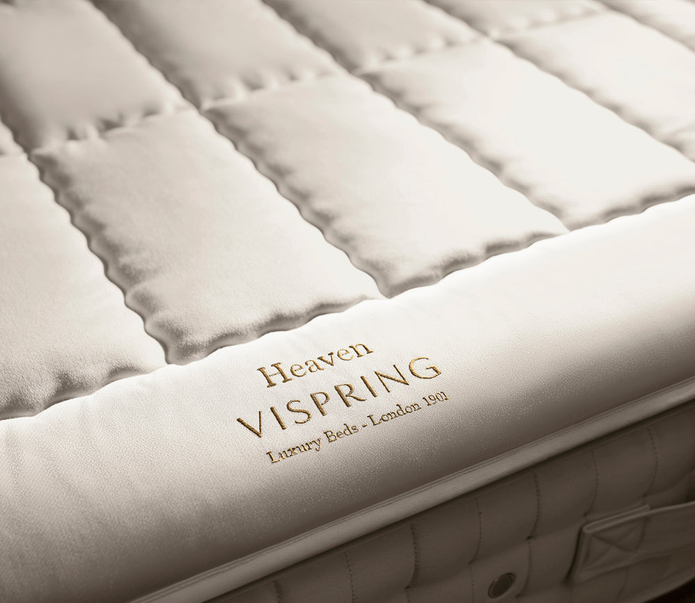 Close-up of Heaven mattress topper in warm off-white/cream fabric, with the words 'Heaven VISPRING Luxury Beds - London 1892' embroidered on the side.
