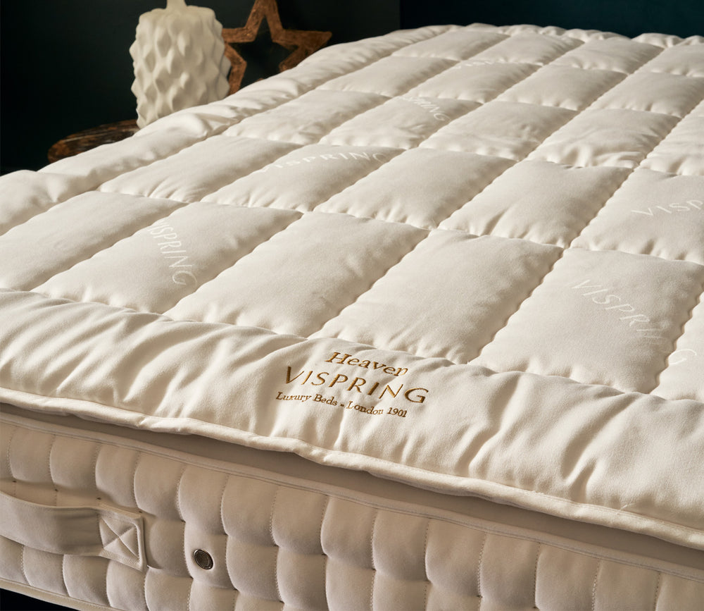 A close-up of Heaven mattress topper in a warm off-white/cream tone, with visible stitching and the brand name 'Heaven Vispring' embroidered on the front.
