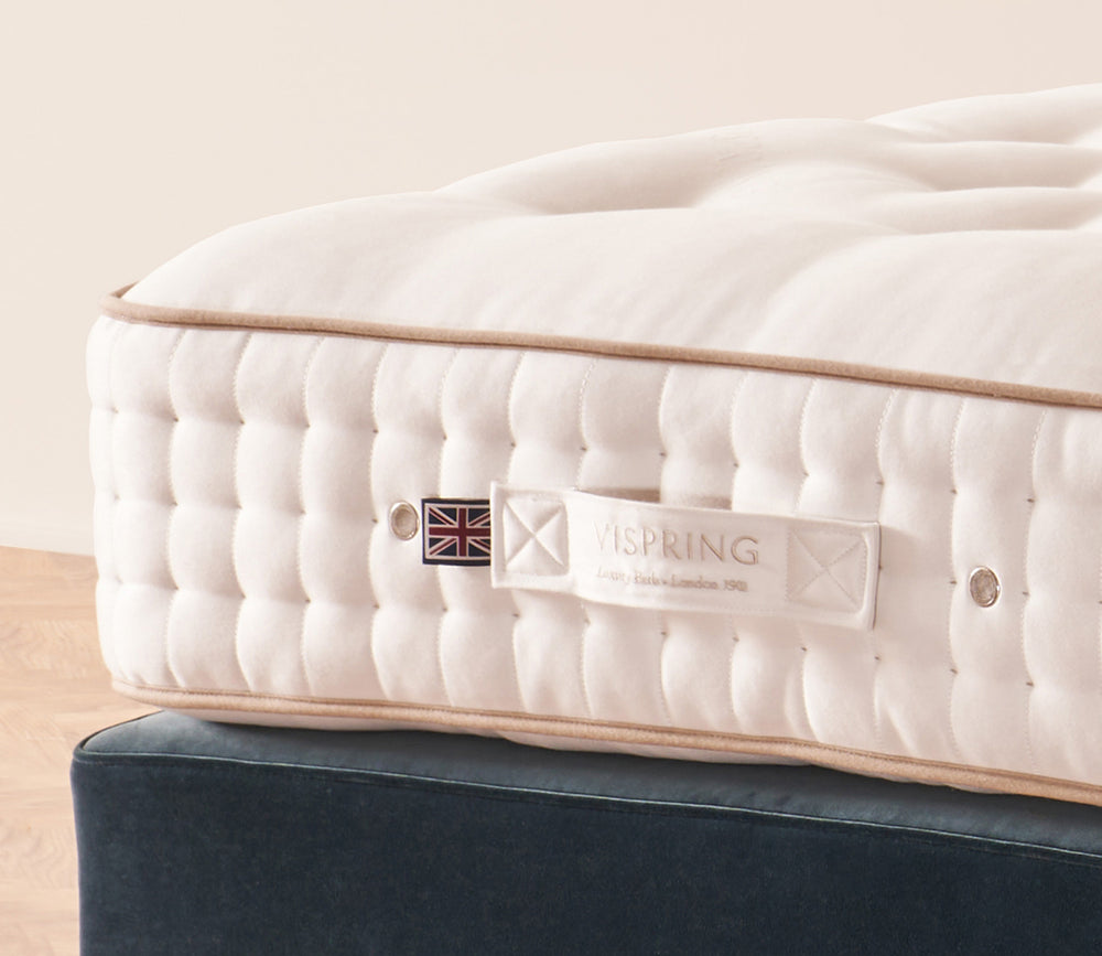 A close-up of Tiara Superb mattress corner with a visible VISPRING label, resting on a dark blue upholstered bed base against a warm beige wall.