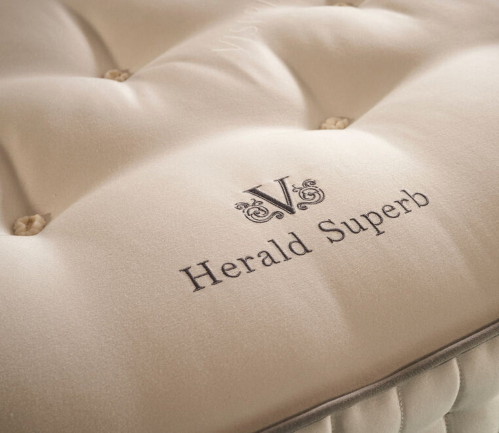 Close-up of a tufted cream-colored mattress or cushion with the embroidered brand name 'Herald Superb' and a monogram logo on the front.
