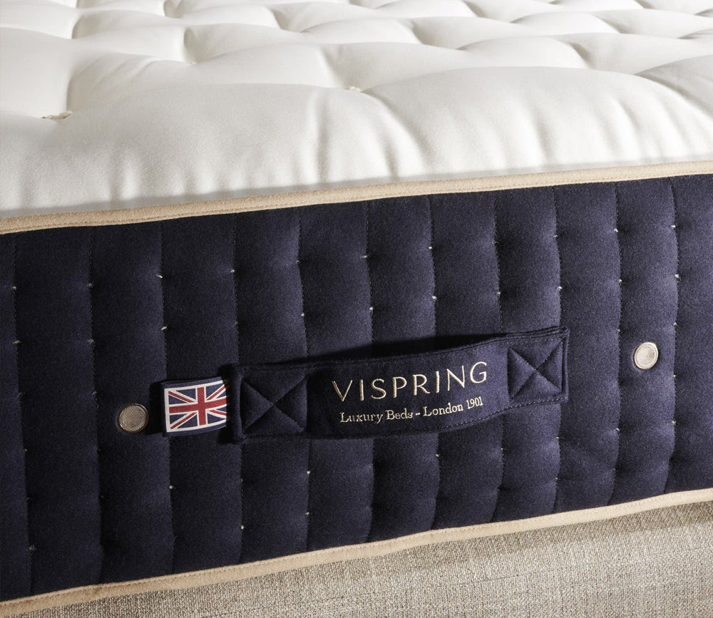 Side view of a Vispring mattress with a hand-tufted white top, deep navy upholstered border, and embroidered Vispring logo.